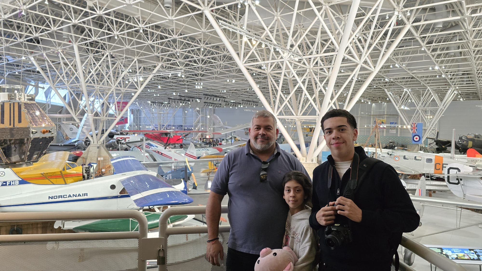 Three people posing in an aviation museum with vintage planes under a white truss ceiling.